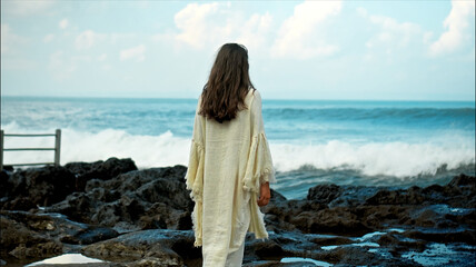 A young European girl with long black hair and a white robe walks to Water blow with cliffs and strong waves of seawater with clouds and blue sea