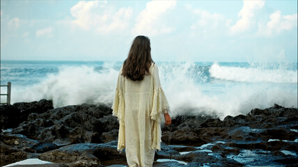 A young European girl with long black hair and a white robe walks to Water blow with cliffs and strong waves of seawater with clouds and blue sea