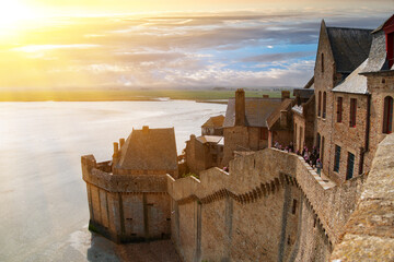 Ancient fortress walls, old houses and buildings of the ancient city-fortress of Mont-Saint-Michel in the rays of the setting sun against the shore of a Atlantic Ocean. Mont Saint Michel, France