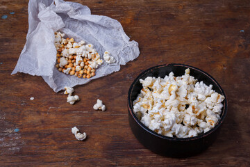 Heap of popcorn in black bowl on wooden rustic background. Some popcorns on white paper