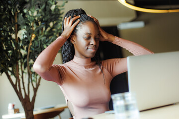 Afro american woman holding hand on head with closed eyes and working online on laptop in coffee house. Black girl working remotely, freelancer, comfortable workplace. Tiredness, overwork, headache.