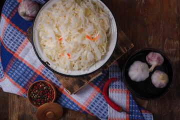 Pickled cabbage in a bowl, garlic and pepper spices on kitchen towel, wooden dark table background. Healthy nutrition, vegan-friendly diet.