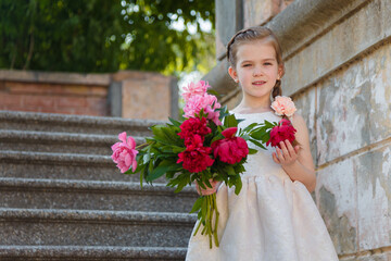 Beautiful little girl in white dress standing on stairs with big peonies bouquet. Spring blossom and carefree childhood concept.