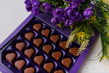 Violet box of heart shaped chocolates, flowers on white background. Valentines day 14 February sweet present