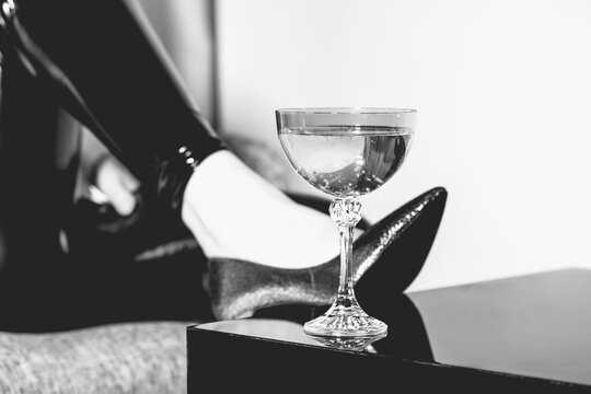 Woman legs with black trousers and fancy high heel shoes placed leg on black mirror table with fancy drink glass on it. Glass is in camera focus. Black and white image