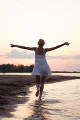 A woman runs along the beach. Photo back view of a beautiful happy blonde in a white summer dress on the river bank against the background of sunset and clouds.