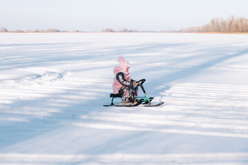 Child rides snowmobile. Little girl in pink warm jacket enjoys walk in nature and sledding on frozen river on sunny winter day. 