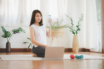 Healthy young Asian woman holding drinking water bottle while using laptop practicing yoga at home