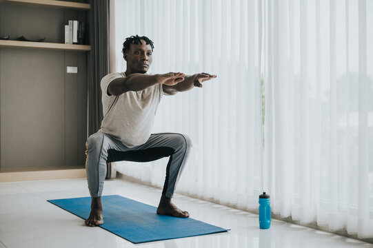 African American man doing squat exercise during workout in livingroom on yoga mat at home