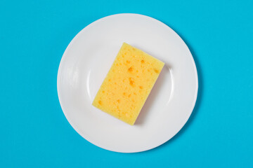 Yellow porous sponge for washing dishes on a white plate on a blue background with copy space. The concept of cleaning, washing dishes and doing housework.