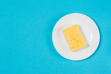 Yellow porous sponge for washing dishes on a white plate on a blue background with copy space. The concept of cleaning, washing dishes and doing housework.