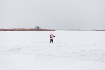 Child on background of winter landscape. Little girl in arm suit and hat with pompom stands on snow-covered ice in middle of lake.