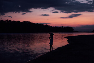 Fototapeta premium The silhouette of a woman in profile against the background of water and sunset. A slender young woman stands in a dress and hat on the beach and looks at the clouds and the picturesque landscape. 