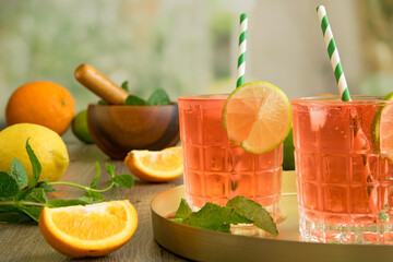 Two glasses full fresh natural lemonade with mint and citrus, standing on tray on wooden table. Cocktail straws sticking up from drink. Slices of orange, fruits on background. Summer refreshing drink