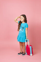 full-length portrait of a long-haired girl carrying a suitcase and looking far away with hand over head, isolated on a pink background.