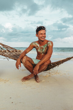 young man sitting on rope hammock at tulum beach in mexico with green body paint art at sunset