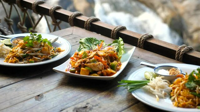 Traditional asian outdoor cafe with thai food with nature waterfall background. Plates with hot fried noodle with prawn pad thai, peanut nuts, green onion and chicken cashew nut on wooden table