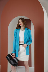 High fashion photo of a beautiful elegant young woman in a pretty aqua blue jacket, blazer, white top, skirt, accessories, black handbag. Brown wall and arch. Studio Shot.