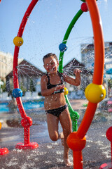 Child is playing in water park. Little girl in black two-piece swimsuit runs on water area of...