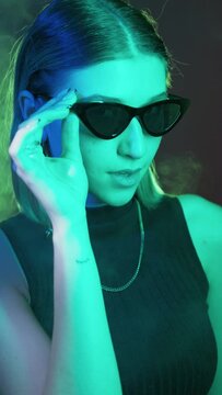 Neon studio portrait. Eyewear fashion. Night life. Stylish attractive woman face winking eye in trendy sunglasses vibrant colorful light smoke vertical.