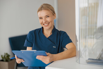 Smiling doctor with clipboard standing near reception in clinic hall and looking camera