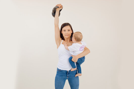 Portrait of young sporty muscular woman with dark hair wearing white top lifting kettle bell, showing muscles biceps.