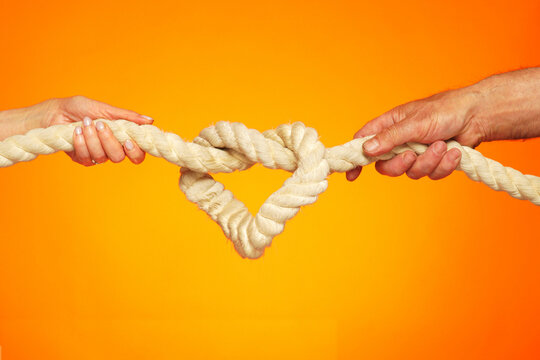 Image of a heart in the form of a rope knot. Male and female hands hold on to a thick rope. Knot tightening. The concept of strong love and inextricable relationship.