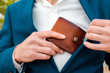 A businessman in a blue jacket and white shirt take off a brown leather wallet in a pocket. Close-up of the hands. The concept of style and fashion - obrazy, fototapety, plakaty