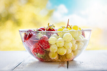 Transparent glass bowl filled with freshly picked strawberries and grapes, washed in water. Blurry...