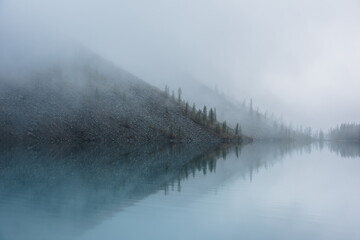 Tranquil meditative misty scenery of glacial lake with pointy fir tops reflection at early morning. Graphic EQ of spruce silhouettes on hill near calm alpine lake in mystery fog. Ghostly mountain lake