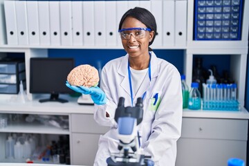 African young woman working at scientist laboratory holding brain looking positive and happy...