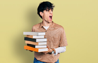 Handsome hipster young man holding a pile of books angry and mad screaming frustrated and furious, shouting with anger. rage and aggressive concept.