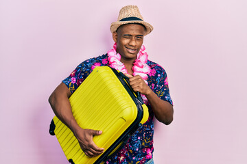 Young black man wearing summer hat holding cabin bag winking looking at the camera with sexy...