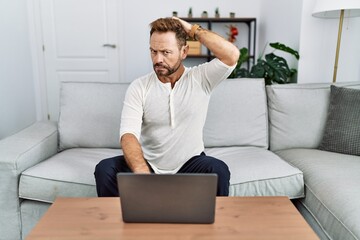 Middle age man using laptop at home confuse and wondering about question. uncertain with doubt, thinking with hand on head. pensive concept.