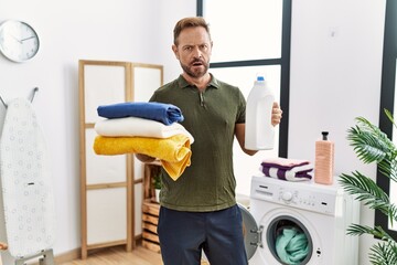 Middle age man holding clean laundry and detergent bottle in shock face, looking skeptical and...