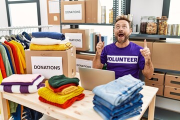 Middle age man wearing volunteer t shirt working with laptop amazed and surprised looking up and pointing with fingers and raised arms.