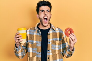 Fototapeta premium Young hispanic man eating doughnut and drinking coffee angry and mad screaming frustrated and furious, shouting with anger looking up.