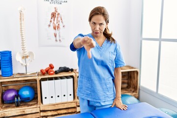 Middle age hispanic physiotherapist woman working at pain recovery clinic looking unhappy and angry showing rejection and negative with thumbs down gesture. bad expression.