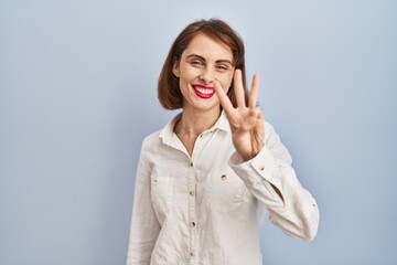 Young beautiful woman standing casual over blue background showing and pointing up with fingers number three while smiling confident and happy.