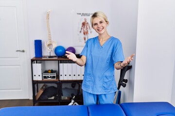 Young physiotherapist woman working at pain recovery clinic smiling cheerful offering hands giving assistance and acceptance.