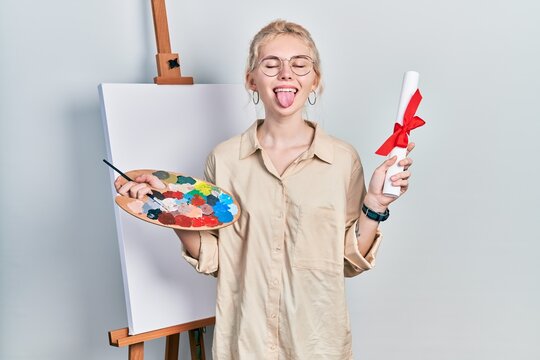 Beautiful caucasian woman with blond hair standing by painter easel stand holding diploma sticking tongue out happy with funny expression.