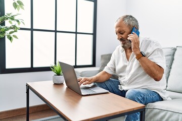 Senior grey-haired man talking on the smartphone using laptop at home