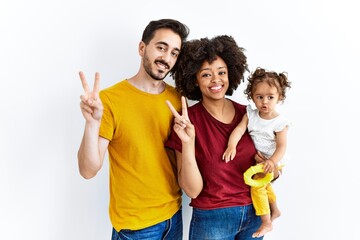 Interracial young family of black mother and hispanic father with daughter smiling looking to the camera showing fingers doing victory sign. number two.