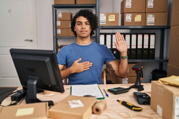 Hispanic man with curly hair working at small business ecommerce swearing with hand on chest and...