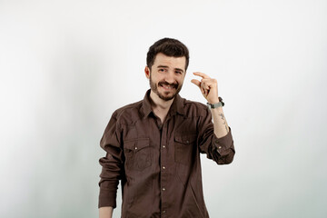 Caucasian man wearing brown shirt posing isolated over white background looking at camera showing measurement with fingers. Showing small size length with a pity.