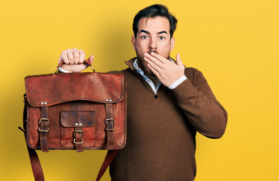 Young hispanic man wearing leather bag covering mouth with hand, shocked and afraid for mistake. surprised expression
