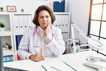 Middle age hispanic woman wearing doctor uniform and stethoscope at the clinic touching mouth with hand with painful expression because of toothache or dental illness on teeth. dentist