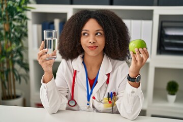 Young african american dietitian woman holding fresh apple and water smiling looking to the side...