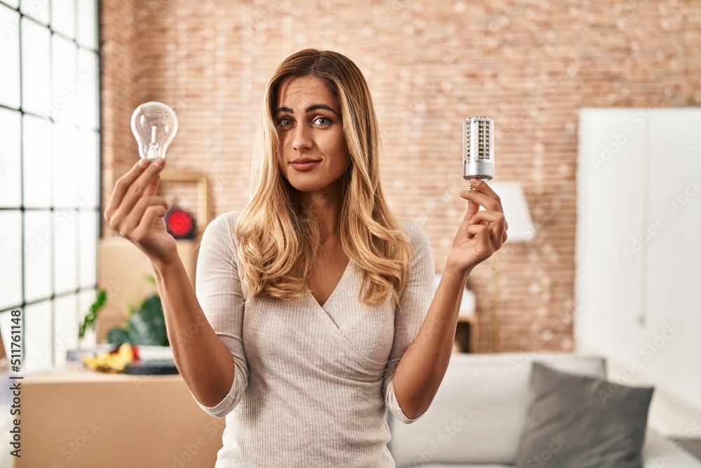 Poster young blonde woman holding led lightbulb and incandescent bulb clueless and confused expression. dou