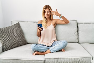 Blonde beautiful young woman sitting on the sofa at home using smartphone shooting and killing...